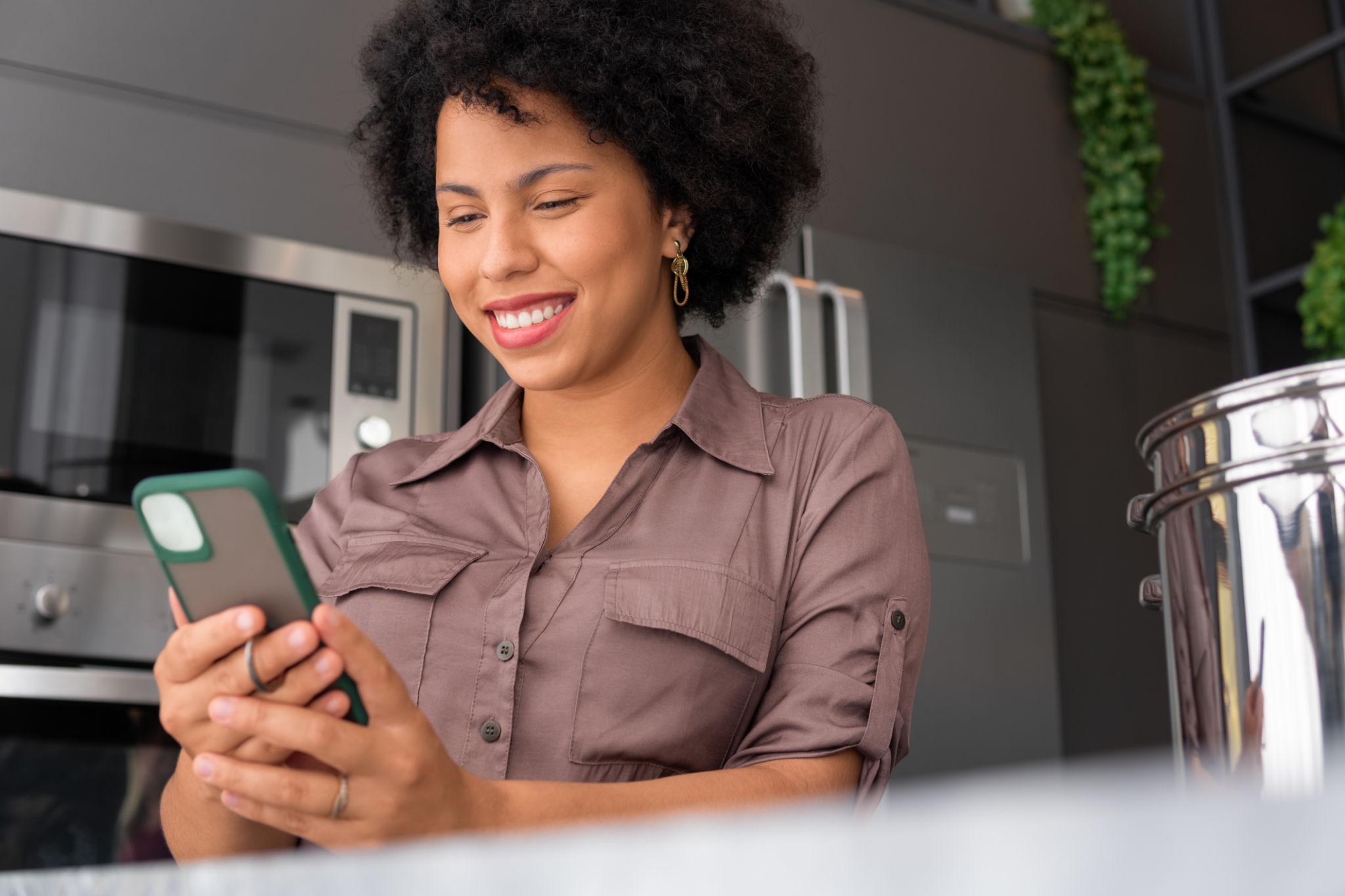Mulher sorrindo enquanto utiliza o celular na cozinha